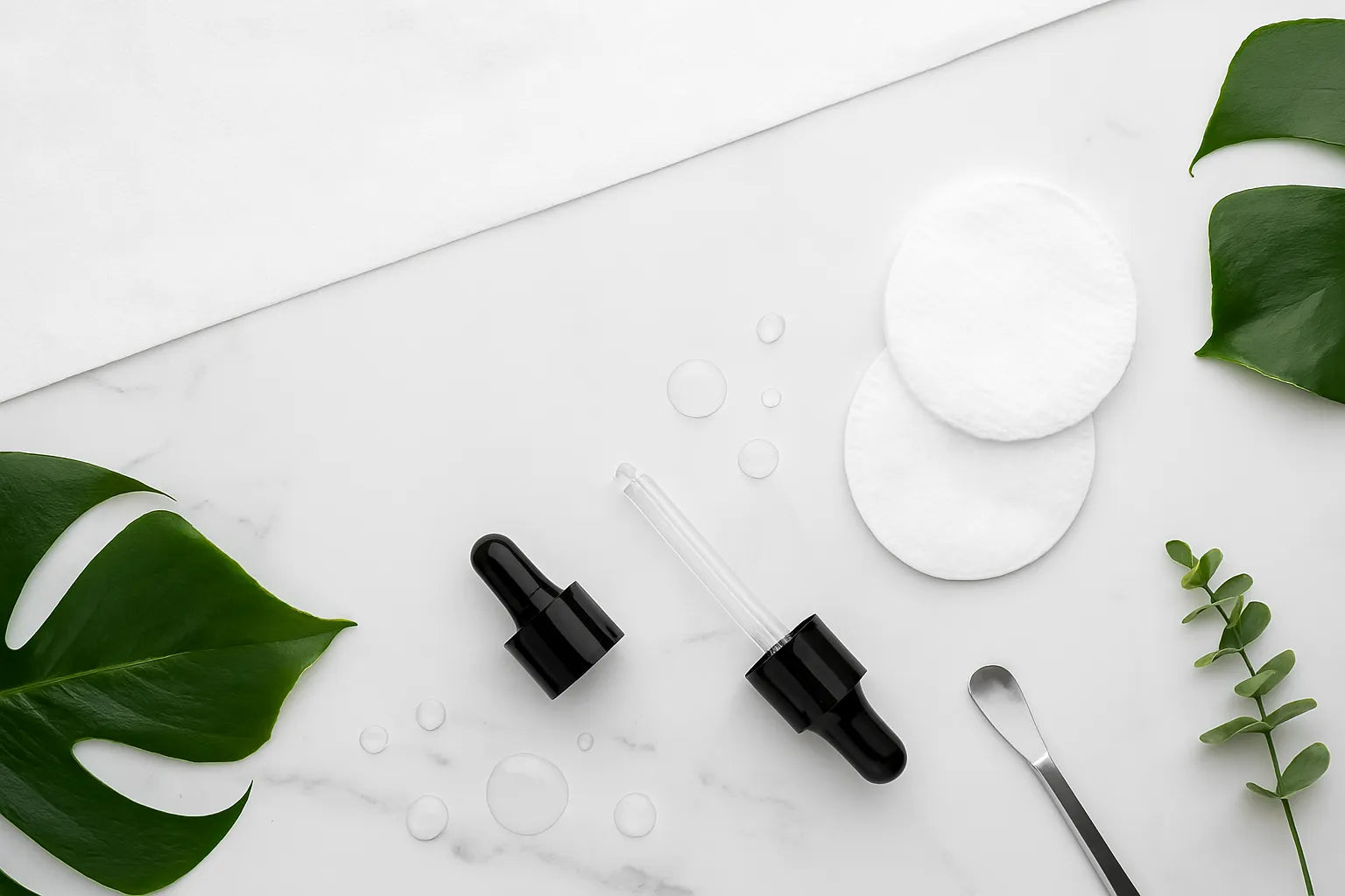 Black serum dropper bottles with cotton pads and cosmetic spatula on white marble background with tropical green leaves